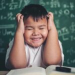 一個小男孩，雙手放在頭上，周圍是書，麵前是一塊寫滿科學符號的黑板，代表準備參加資格考試